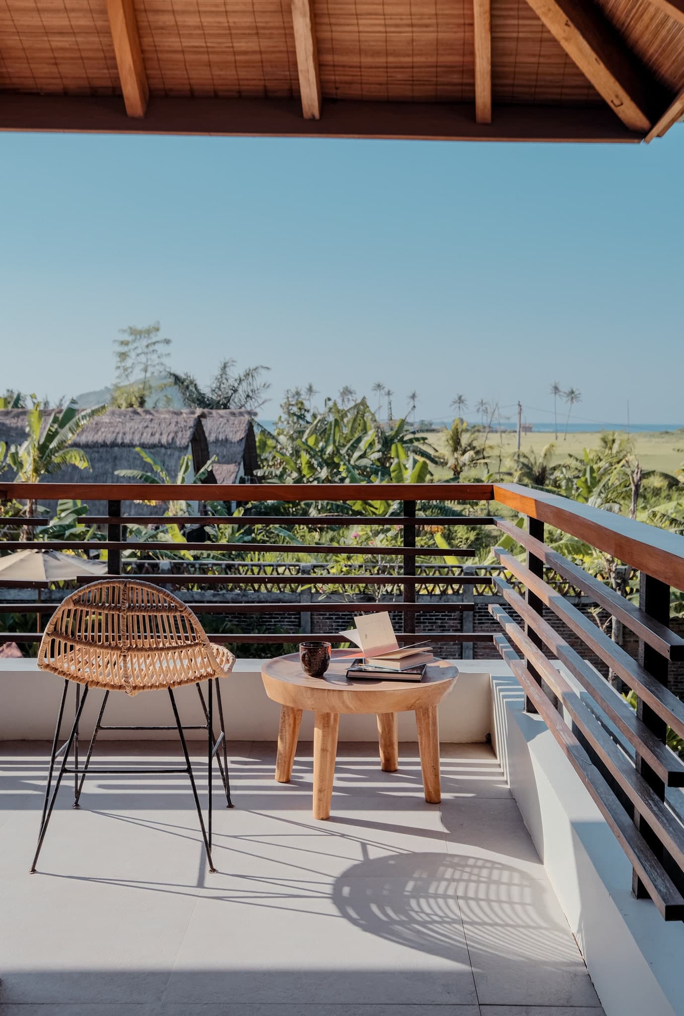 Private balcony at Villa Gudea, Lombok – Featuring a rattan chair and wooden table overlooking tropical gardens and ocean views, perfect for relaxing with a book and coffee