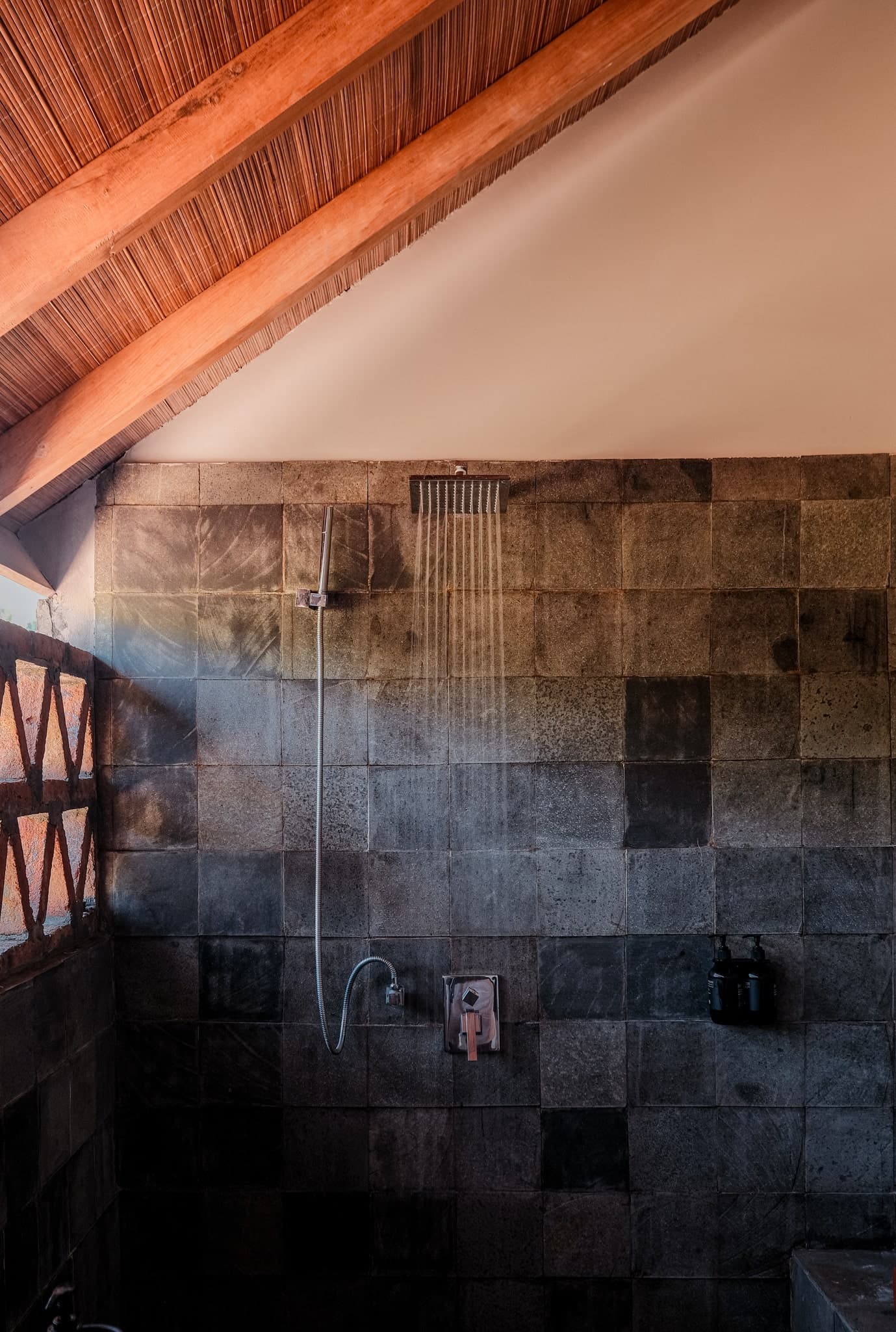 Rustic outdoor shower at Villa Gudea, Lombok — Featuring dark stone tiles, a rainfall showerhead, and natural wood accents under a sloped bamboo ceiling for a tropical, nature-inspired bathing experience