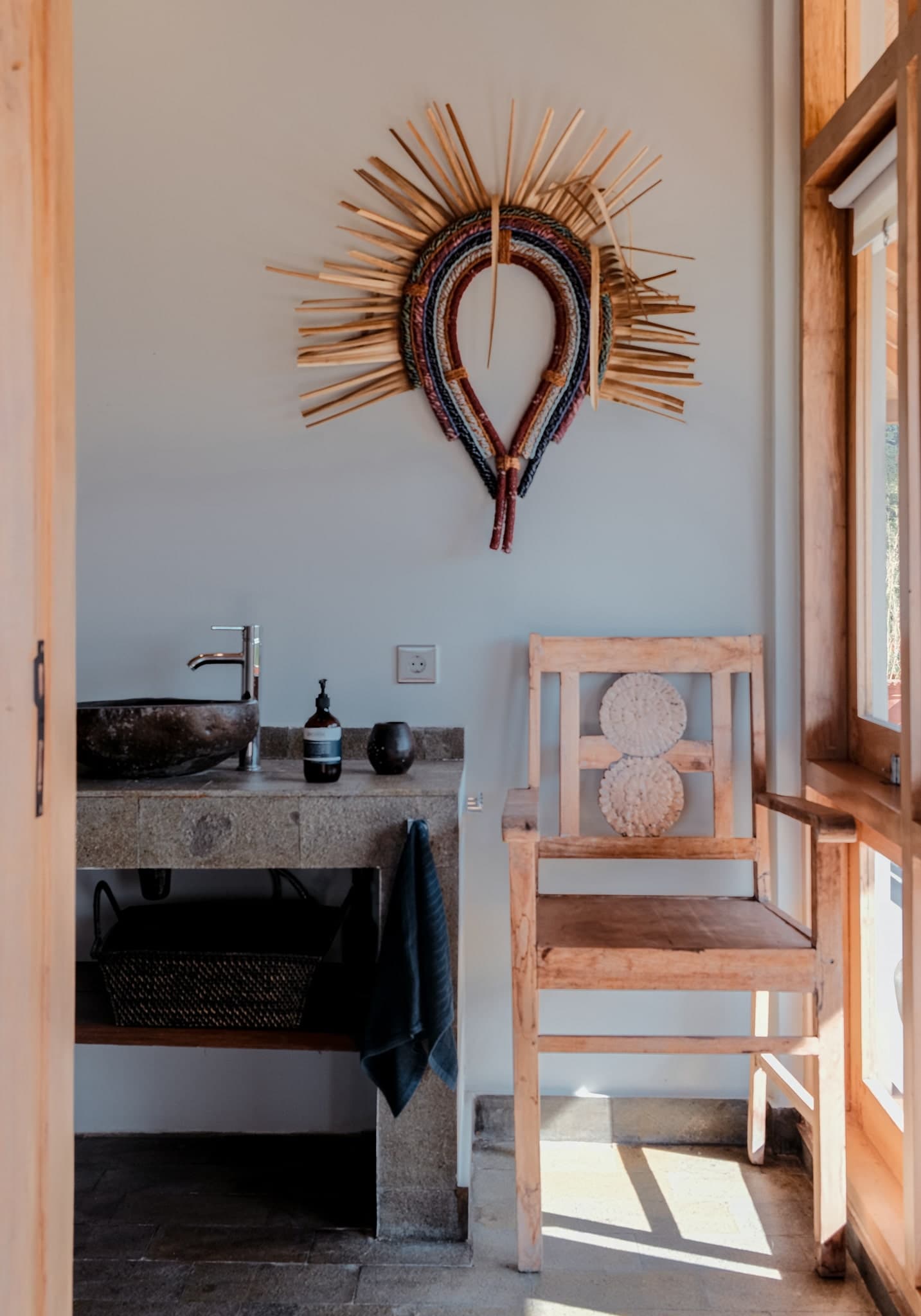 Rustic bathroom corner at Villa Gudea, Lombok – Featuring a stone sink, handmade wooden chair, and vibrant Indonesian wall art with natural light streaming through large windows