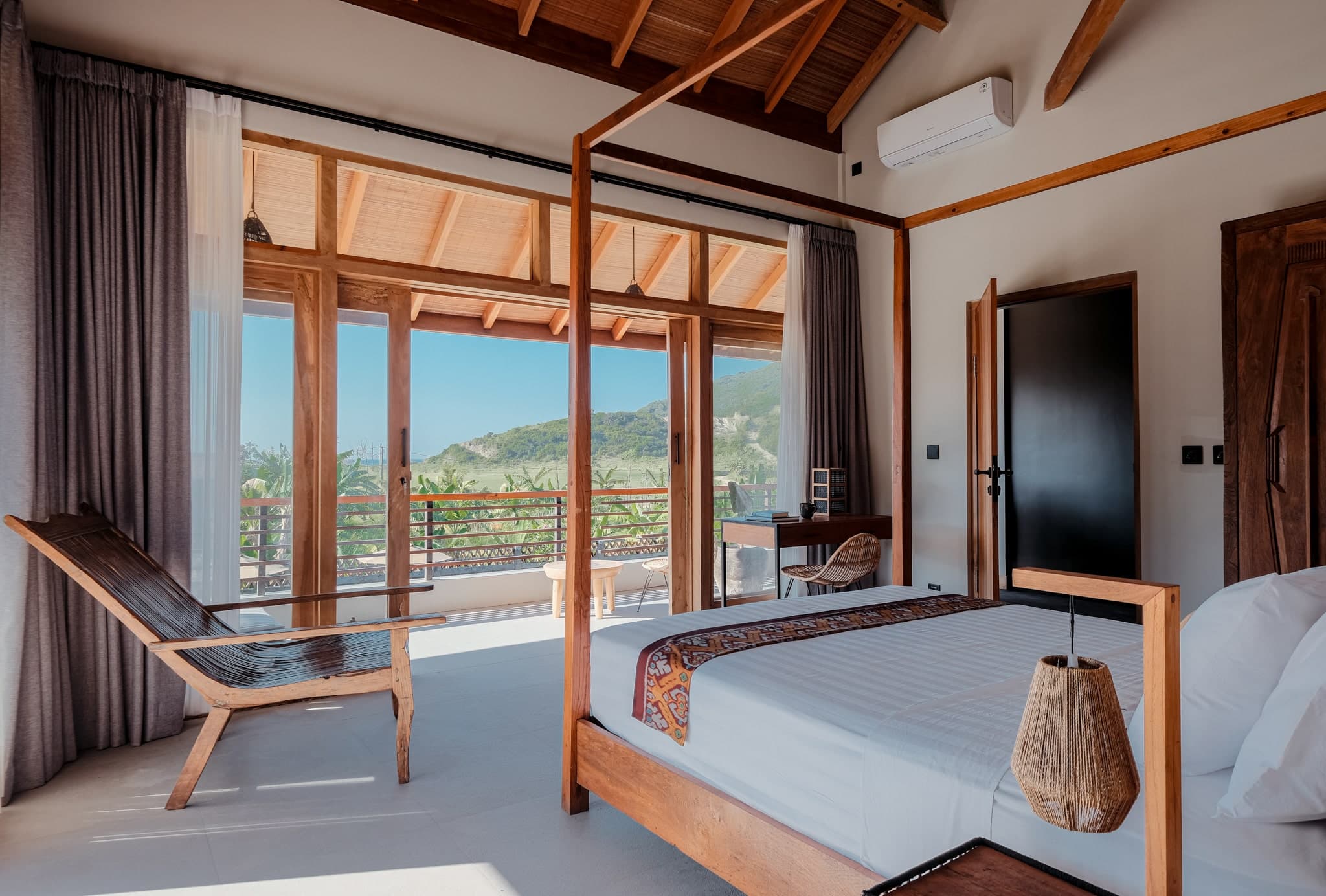 Spacious bedroom at Villa Gudea, Lombok – Showcasing a wooden four-poster bed, handcrafted furniture, and floor-to-ceiling windows with panoramic views of lush hills and tropical landscapes