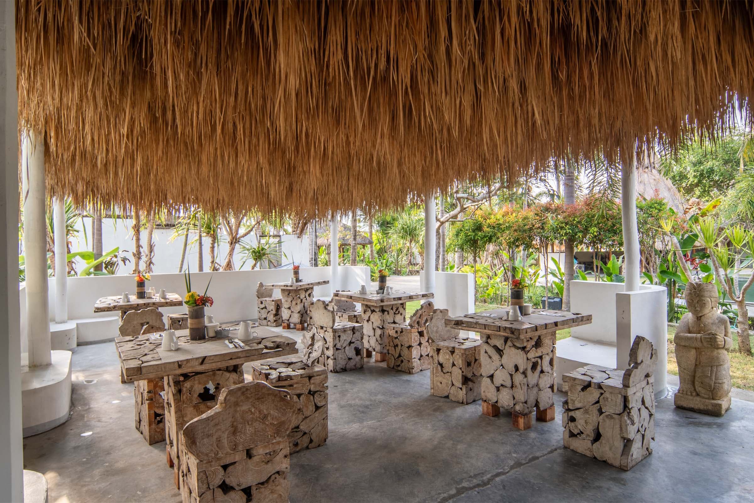 Open-air dining area at Tiki Lodge with rustic wooden tables and chairs made from driftwood, under a thatched roof, surrounded by lush tropical gardens and stone statues