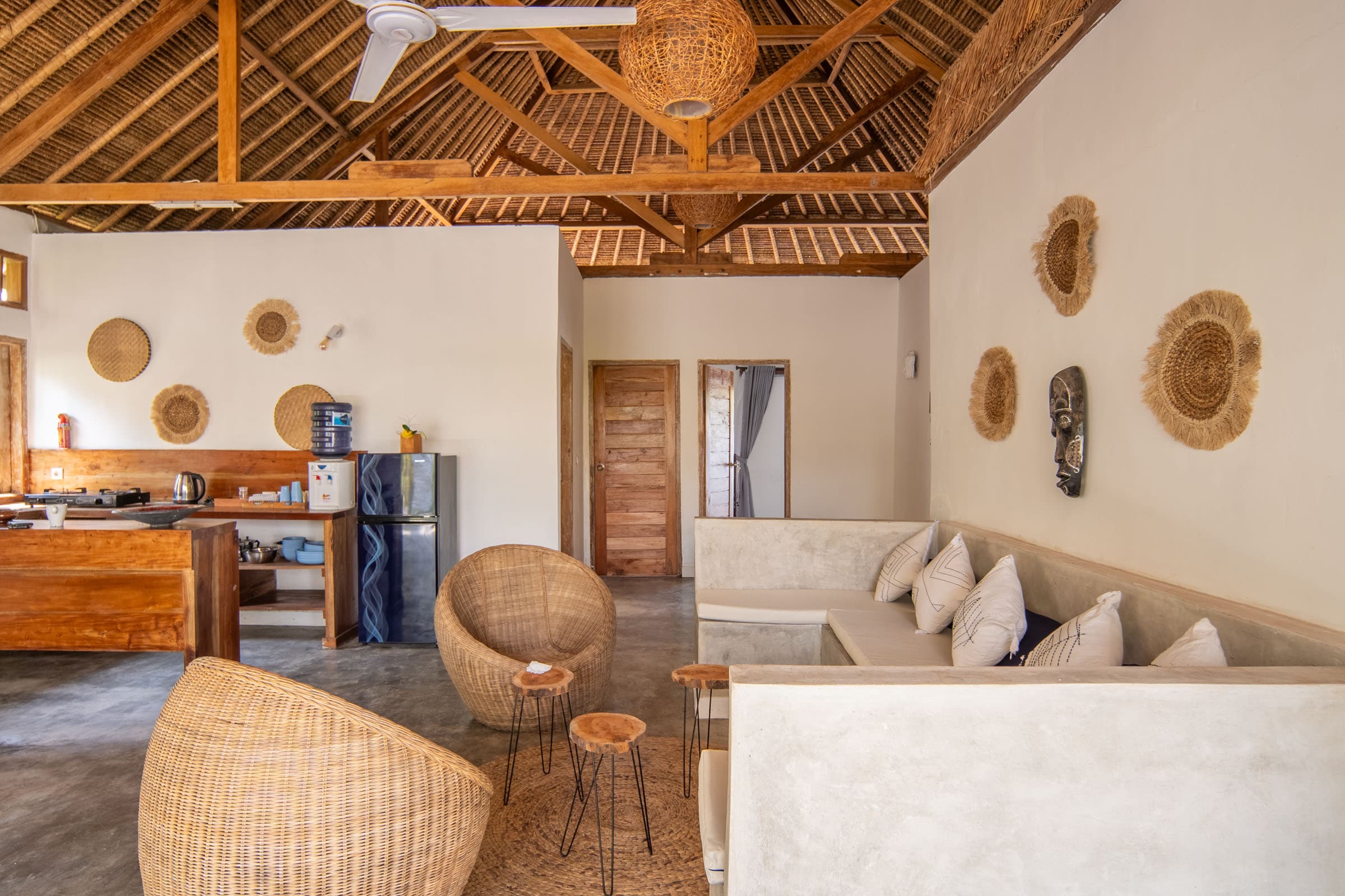 Cozy living area at Tiki Lodge with rattan chairs, a built-in sofa with white cushions, and a wooden kitchen, featuring rustic decor, woven wall hangings, and an open-beam thatched ceiling