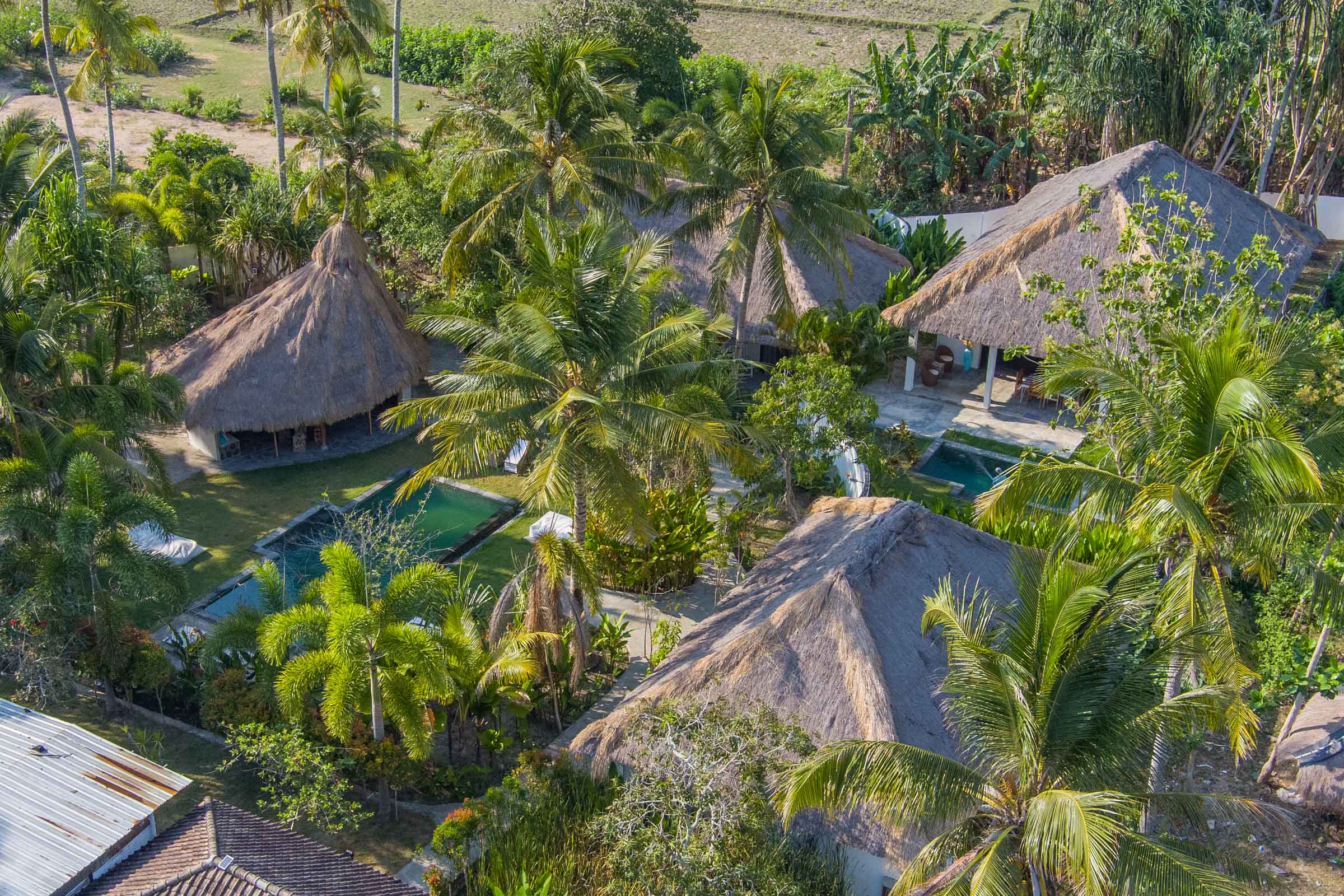 Aerial view of Tiki Lodge nestled amidst lush greenery in Selong Belanak, Lombok, with traditional thatched-roof bungalows surrounding a central swimming pool